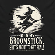 A black long-sleeve hoodie with a white graphic print of a witch riding a broomstick in front of a full moon, with the text 'HOLD MY BROOMSTICK. SHIT'S ABOUT TO GET REAL!' printed below, shown on a plain white background.