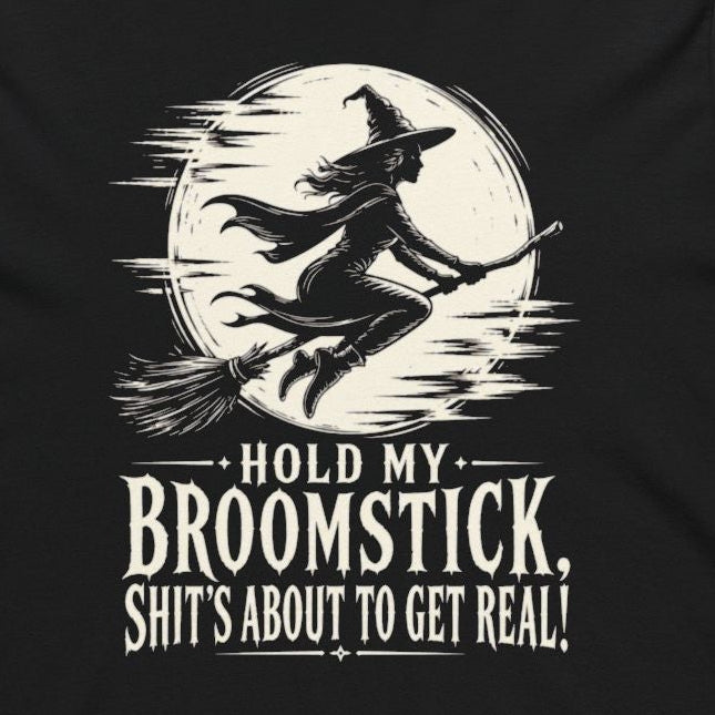 A black long-sleeve hoodie with a white graphic print of a witch riding a broomstick in front of a full moon, with the text 'HOLD MY BROOMSTICK. SHIT'S ABOUT TO GET REAL!' printed below, shown on a plain white background.