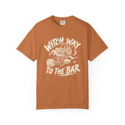 A rust-orange short-sleeve t-shirt with a white graphic print on the front that reads 'WITCH WAY TO THE BAR' around an illustration of a witch riding a broomstick.