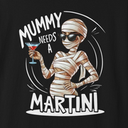 A black short-sleeve crewneck t-shirt featuring a cartoon graphic of a mummy holding a martini glass, with the text 'MUMMY NEEDS A MARTINI' printed in white and beige.