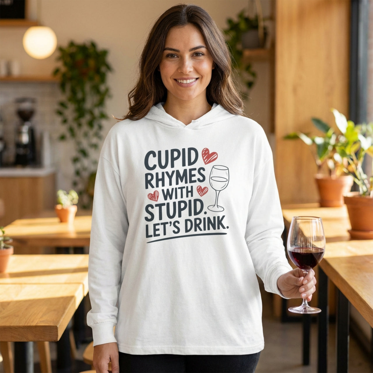 brunette model wearing white featherweight hoodie with the phrase "Cupid Rhymes With Stupid Let's Drink" with a minimalist sketched wine glass and valentine's hearts to complete the design

