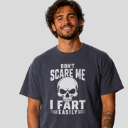 A man wearing a dark navy blue graphic t-shirt with a white skull and the text 'DON'T SCARE ME I FART EASILY' printed on the front, set against a solid light gray background.