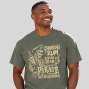 A man wearing an olive green graphic t-shirt with a pirate-themed design and text that reads 'DRINKING RUM BEFORE 10 AM MAKES YOU A PIRATE NOT AN ALCOHOLIC'.