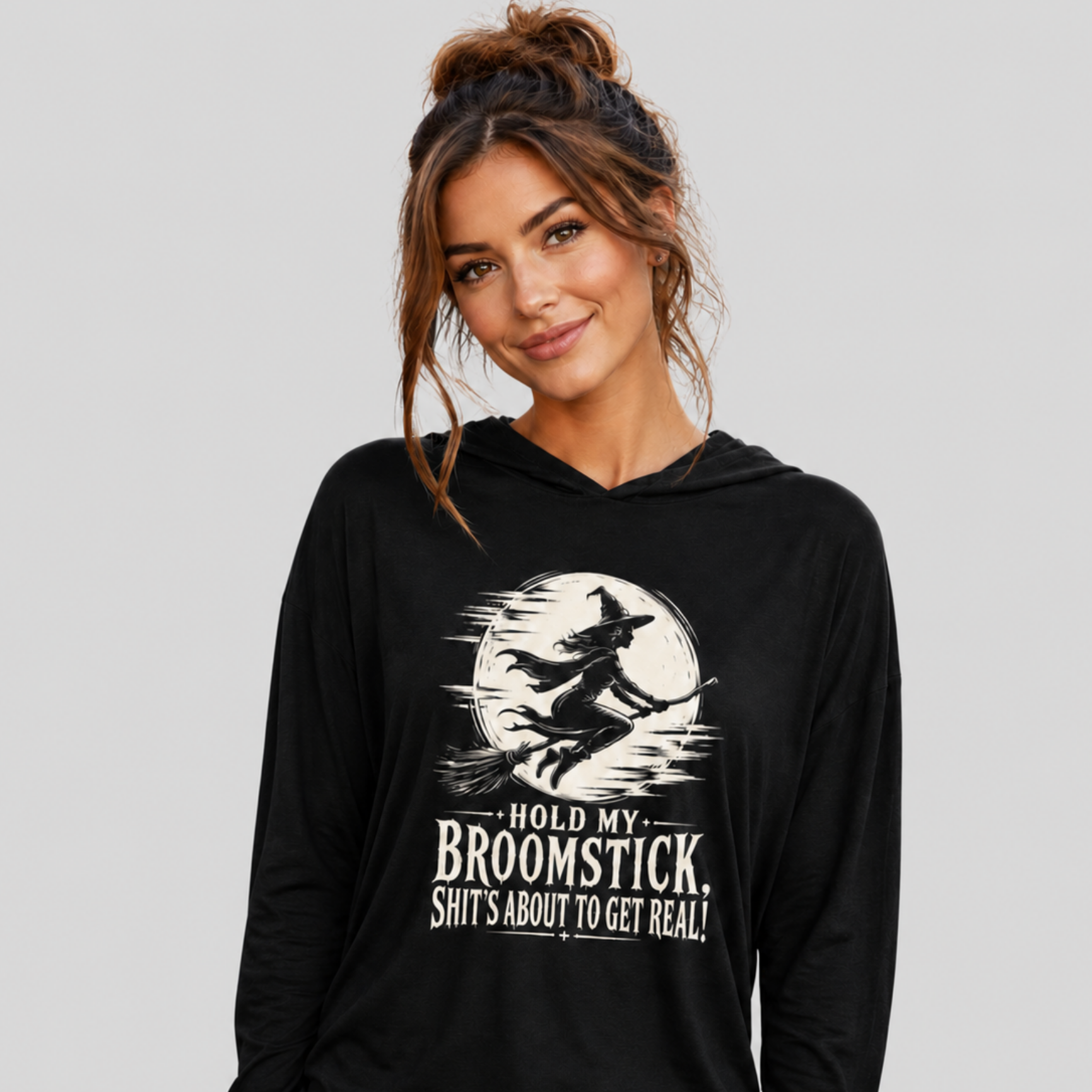 A woman wearing a black long-sleeve hoodie with a white graphic of a witch riding a broomstick in front of a full moon, with the text 'HOLD MY BROOMSTICK SHIT'S ABOUT TO GET REAL!'.
