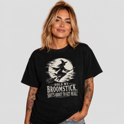 A woman with blonde hair and tattoos wearing a black graphic t-shirt with a white print of a witch riding a broomstick in front of a full moon and the text 'HOLD MY BROOMSTICK, SHIT'S ABOUT TO GET REAL!'.