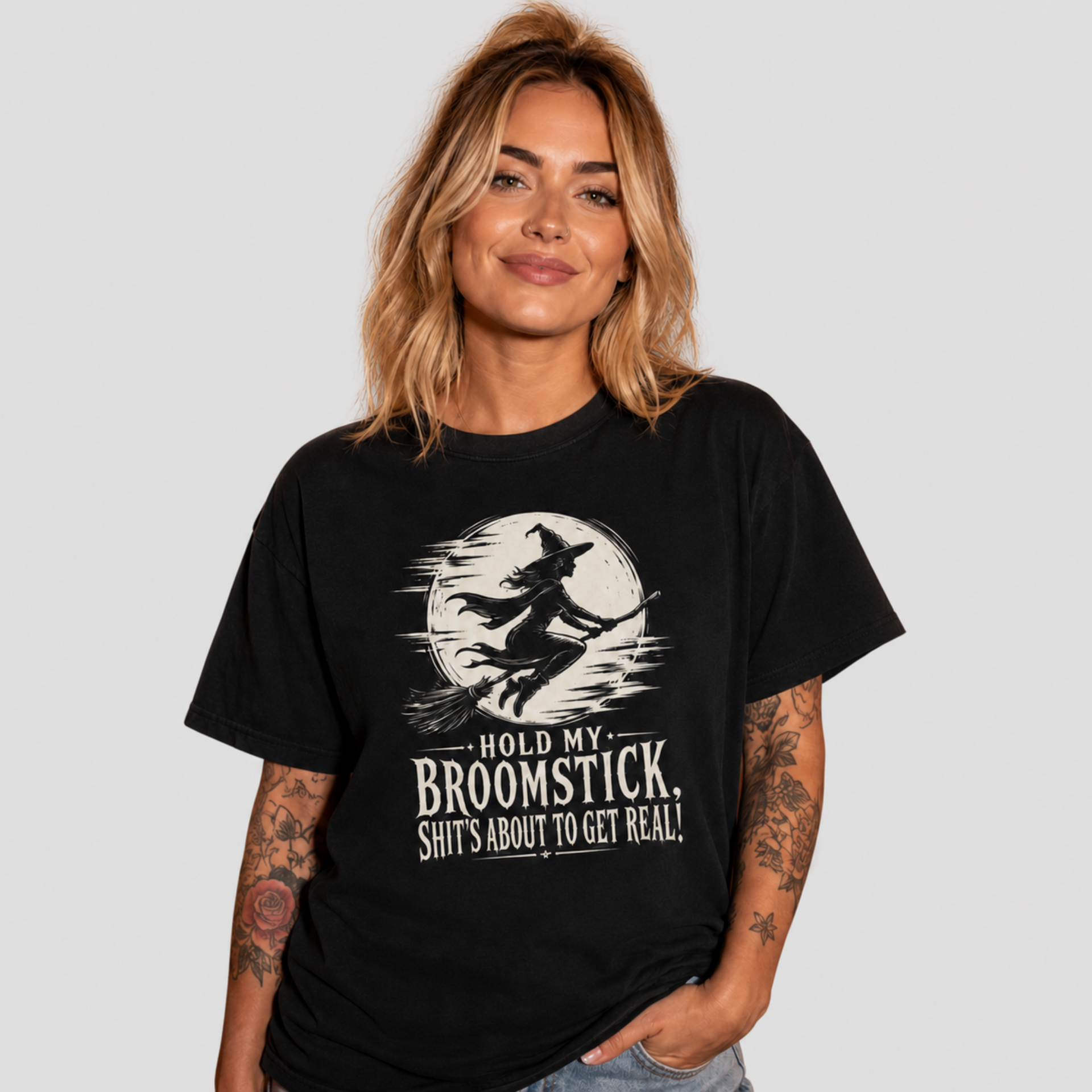 A woman with blonde hair and tattoos wearing a black graphic t-shirt with a white print of a witch riding a broomstick in front of a full moon and the text 'HOLD MY BROOMSTICK, SHIT'S ABOUT TO GET REAL!'.
