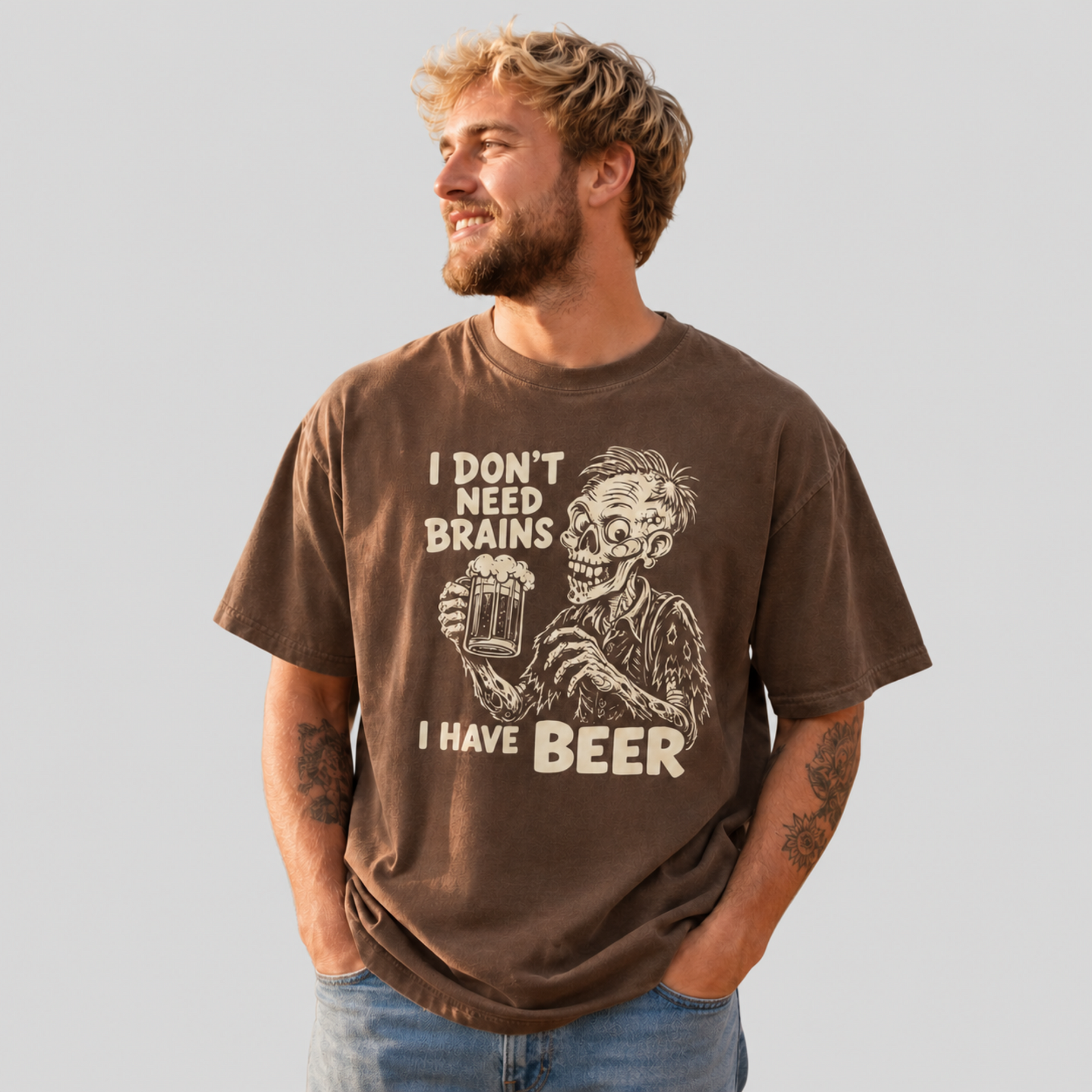 A man wearing a brown graphic t-shirt with a skeleton holding a beer mug and the text 'I DON'T NEED BRAINS I HAVE BEER', paired with blue jeans, set against a solid light gray background.