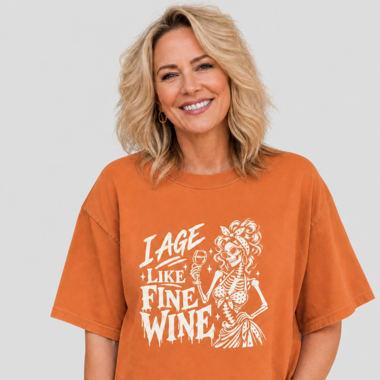 A woman wearing an oversized burnt orange graphic t-shirt with a white distressed print that reads 'I AGE LIKE FINE WINE' next to a skeleton holding a glass of wine, set against a solid light gray background.