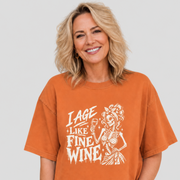 A woman wearing an oversized burnt orange graphic t-shirt with a white distressed print that reads 'I AGE LIKE FINE WINE' next to a skeleton holding a glass of wine, set against a solid light gray background.