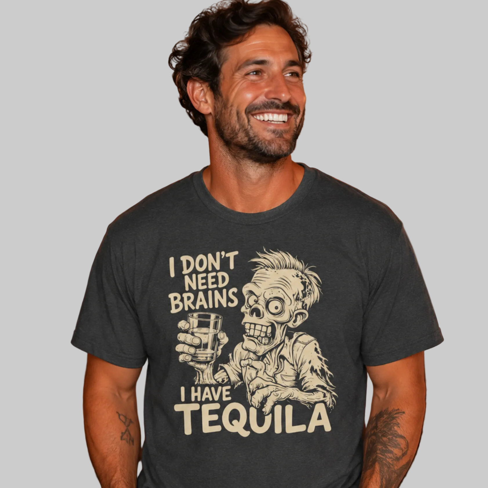 A man wearing a dark charcoal gray graphic t-shirt with a zombie holding a tequila glass and the text 'I DON'T NEED BRAINS I HAVE TEQUILA'. The background is a cozy, warmly lit Halloween-themed room with string lights, pumpkins, and spooky decorations.