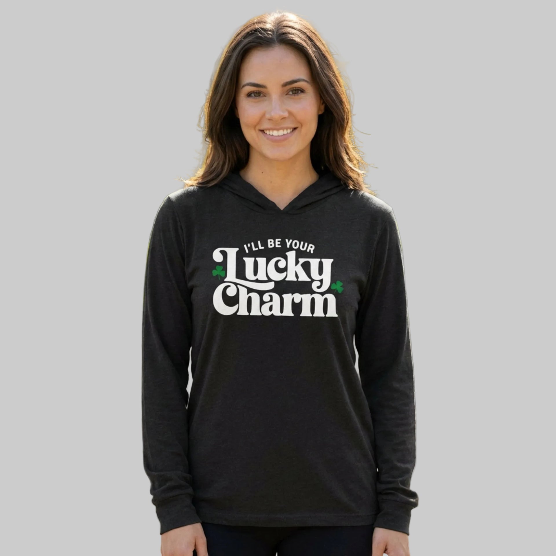 brunette woman wearing a black lightweight St Patricks  Day hoodie with the Phrase “ill be your lucky charm”  with  shamrocks to complete the design