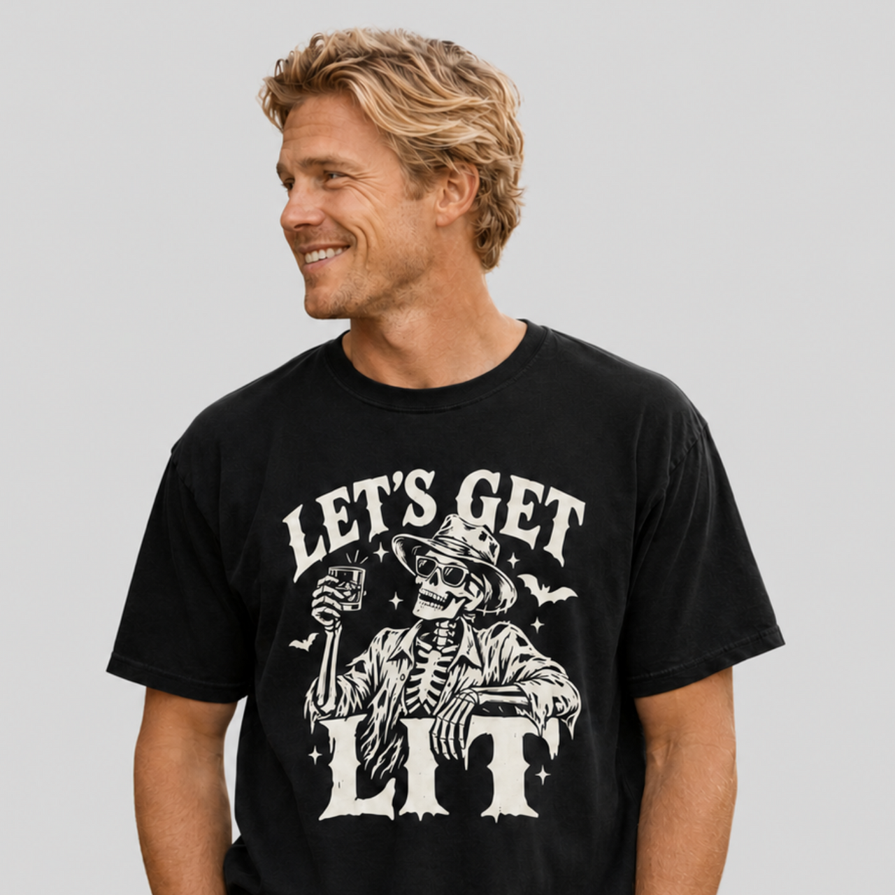 A man wearing a black graphic t-shirt with a white skeleton and 'LET'S GET LIT' print, paired with khaki pants, against a solid gray background.