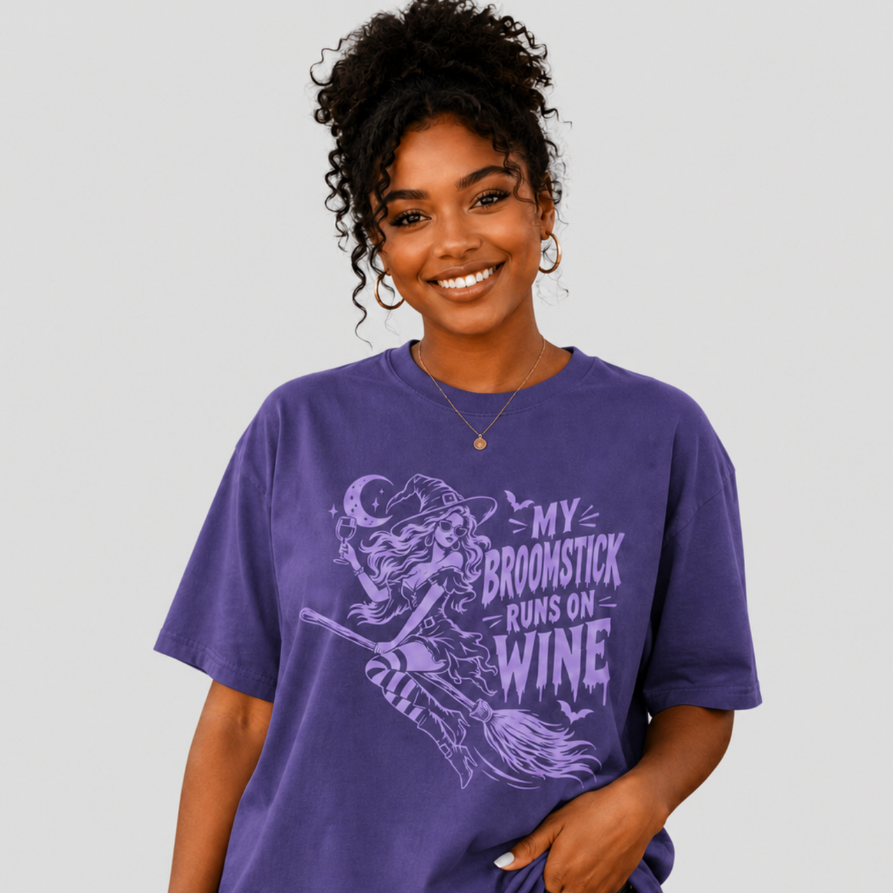 A woman wearing a purple graphic t-shirt with a light purple witch riding a broomstick design and the text 'MY BROOMSTICK RUNS ON WINE'. She is posing against a solid light gray background.
