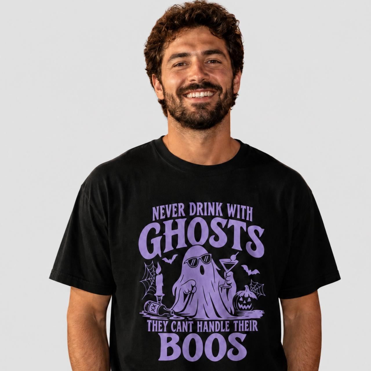 A smiling man wearing a black graphic t-shirt with a purple Halloween-themed design that reads 'NEVER DRINK WITH GHOSTS THEY CAN'T HANDLE THEIR BOOS' around an illustration of a ghost, bats, and a pumpkin.
