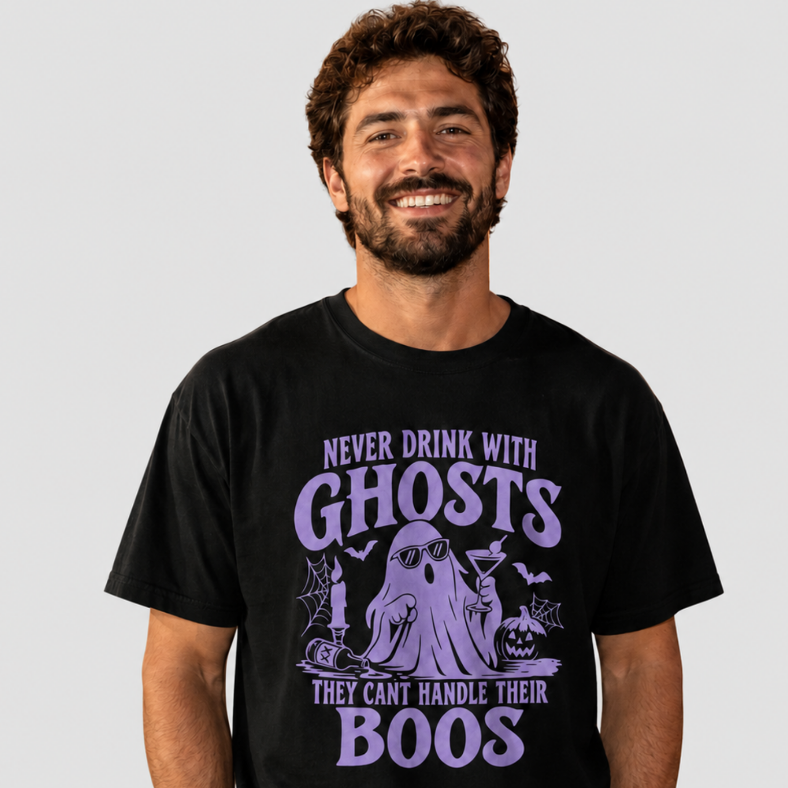 A smiling man wearing a black graphic t-shirt with a purple Halloween-themed design that reads 'NEVER DRINK WITH GHOSTS THEY CAN'T HANDLE THEIR BOOS' around an illustration of a ghost, bats, and a pumpkin.