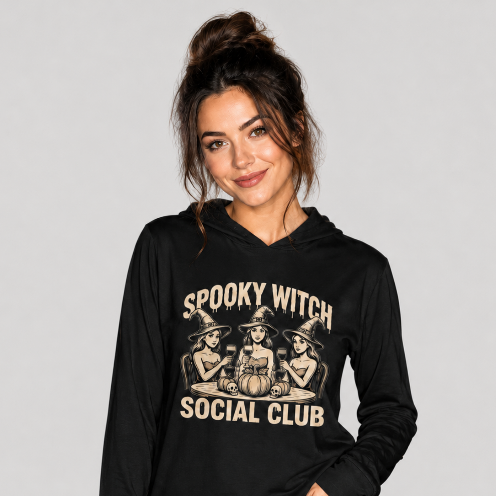 A woman wearing a black long-sleeve hoodie with a beige graphic that reads 'Spooky Witch Social Club' and shows three illustrated witches sitting at a table, paired with distressed blue jeans against a light gray studio background.