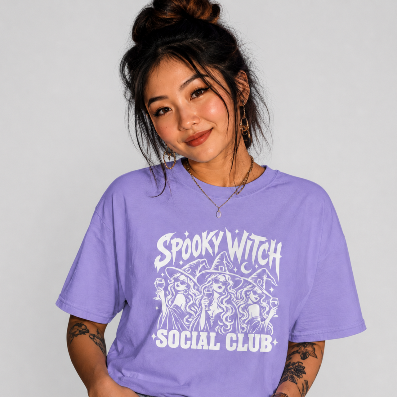 A woman wearing a lavender purple graphic t-shirt with a white 'Spooky Witch Social Club' print, paired with blue jeans, against a light gray studio background.