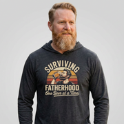 Man wearing a heather navy lightweight hoodie with the phrase 'Surviving Fatherhood One Beer at a Time' funny Father's day gift for dad