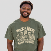 A man wearing an olive green graphic t-shirt with a beige 'Trick or Treat Give Me Something Good to Drink' Halloween print, paired with light blue jeans, against a solid light gray background.