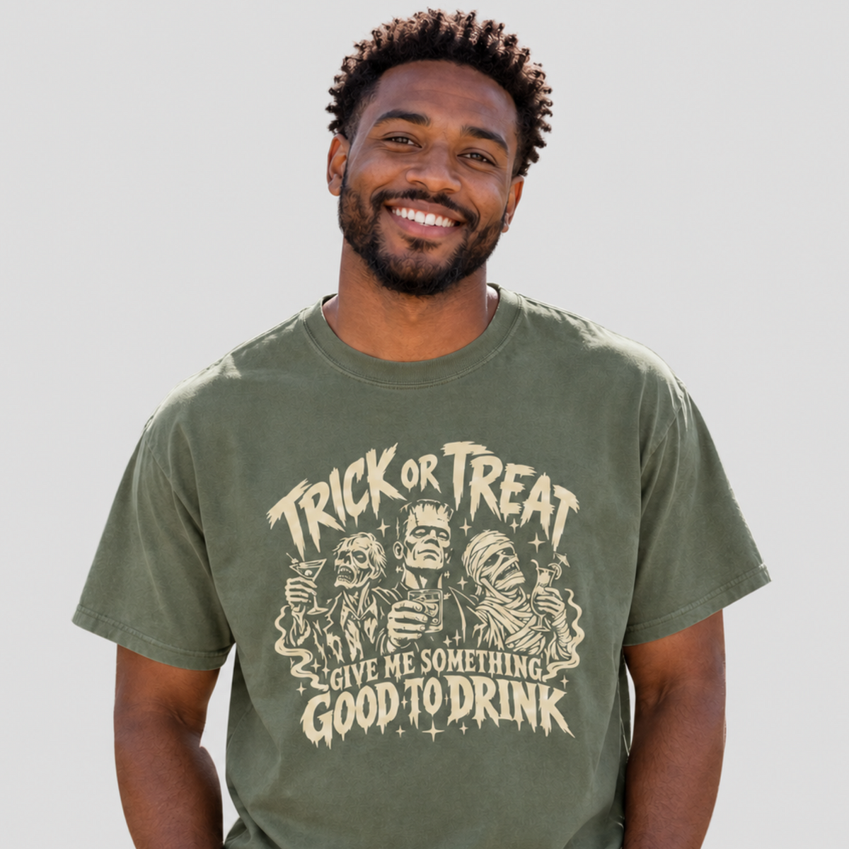 A man wearing an olive green graphic t-shirt with a beige 'Trick or Treat Give Me Something Good to Drink' Halloween print, paired with light blue jeans, against a solid light gray background.