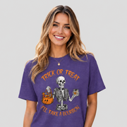 A woman wearing a purple short-sleeve graphic t-shirt with a Halloween design featuring a skeleton, a jack-o'-lantern, and orange text that reads 'TRICK OR TREAT I'LL TAKE A BOURBON'.
