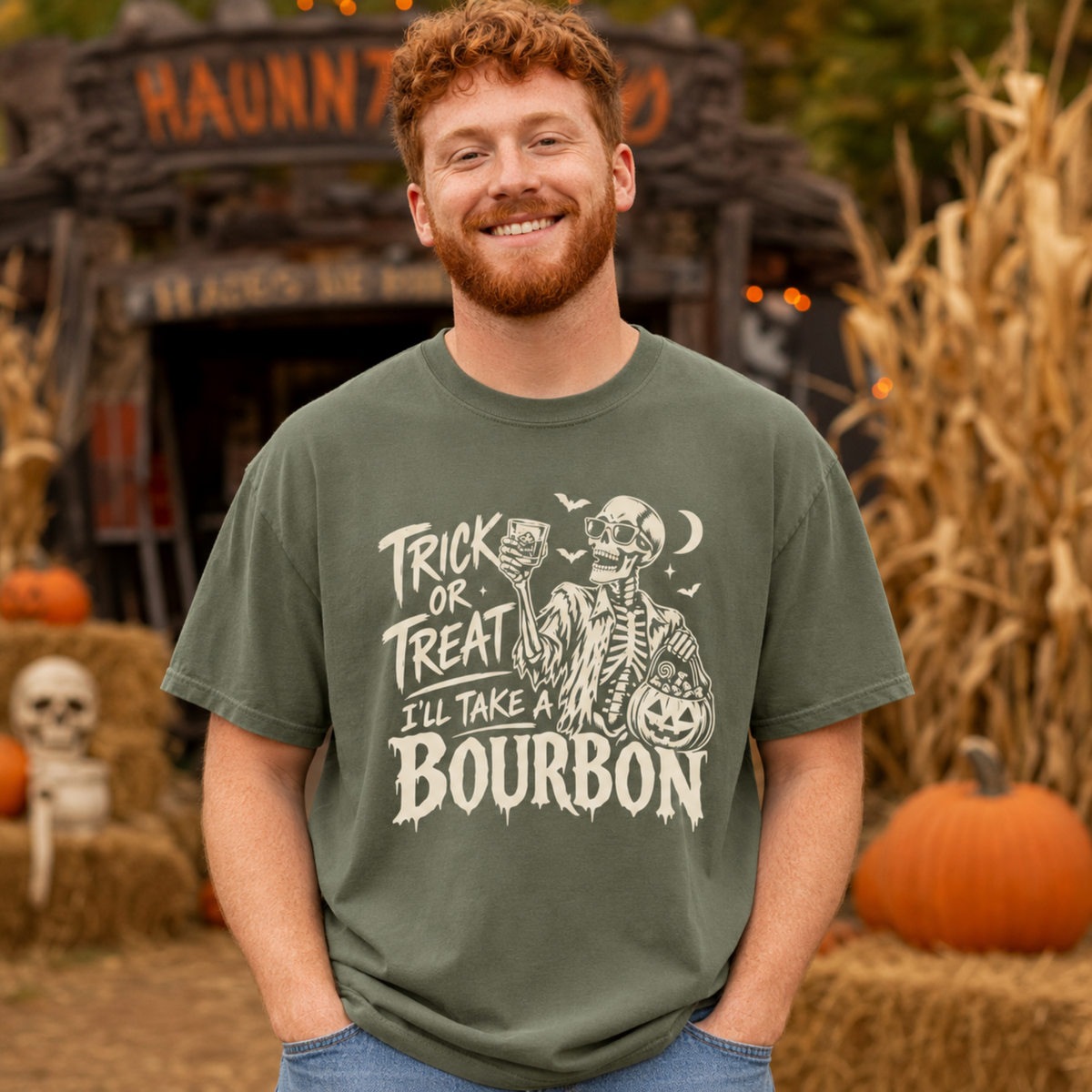 A smiling man with red hair and a beard stands outdoors wearing an olive green graphic T-shirt with a white skeleton and text reading 'TRICK OR TREAT I'LL TAKE BOURBON.' The background shows a Halloween-themed rustic sign, pumpkins, hay, and dry cornstalks.