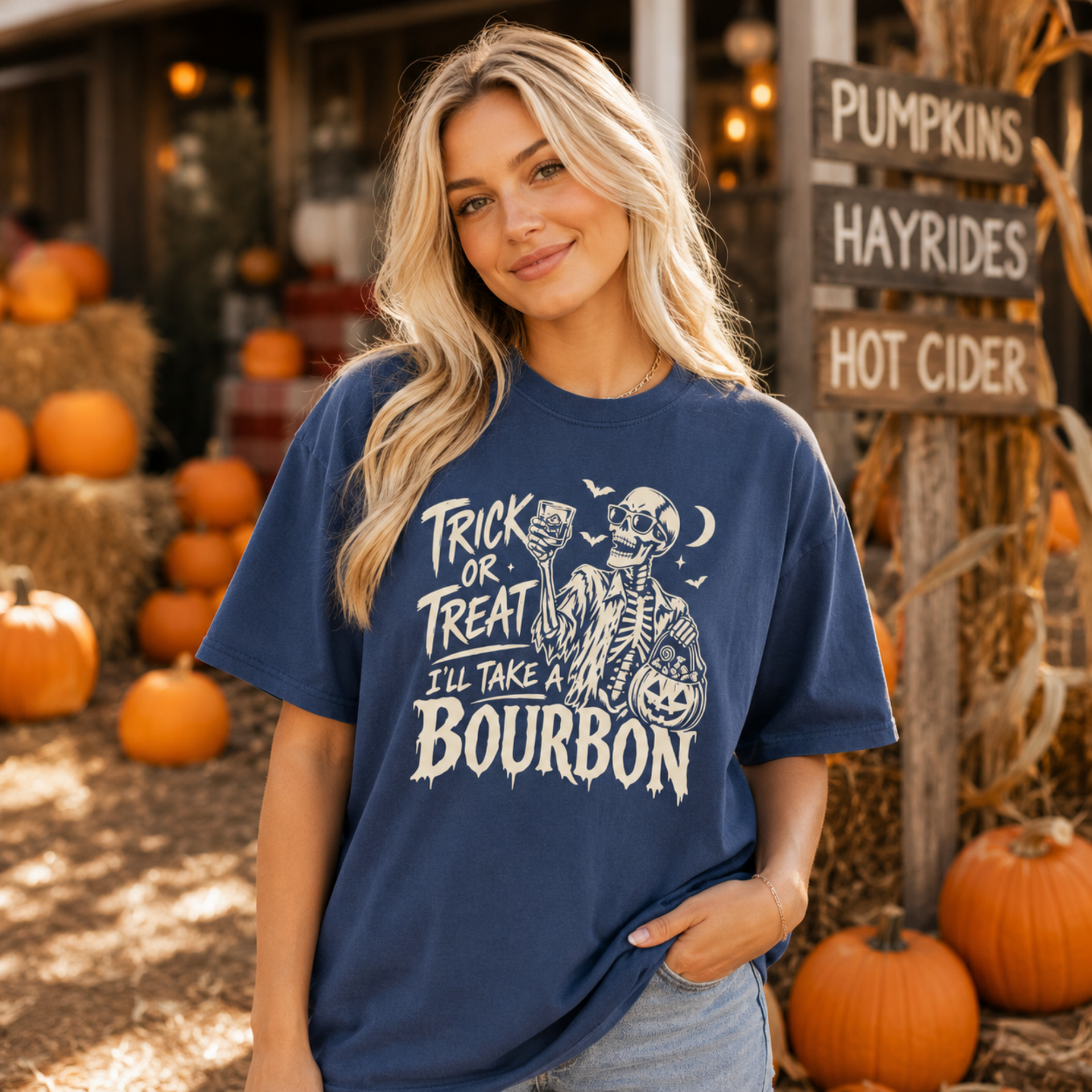 A woman wearing a navy blue graphic t-shirt with a white skeleton and text reading 'TRICK OR TREAT I'LL TAKE A BOURBON' in a rustic autumn setting with pumpkins and a 'Pumpkins Hayrides Hot Cider' sign.