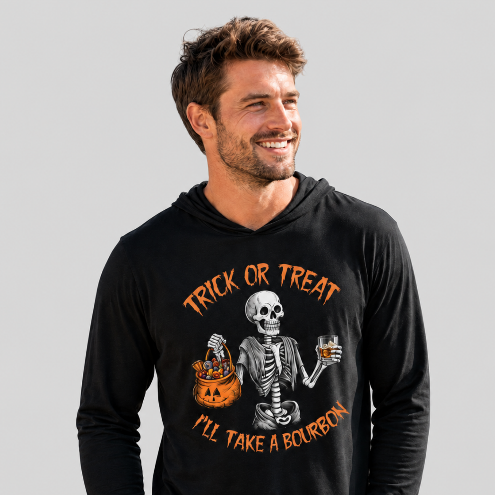 A man wearing a black long-sleeve hoodie with a Halloween graphic of a skeleton holding an orange jack-o'-lantern and a glass of amber liquor, with the text 'TRICK OR TREAT I'LL TAKE A BOURBON' in orange, against a solid gray background.
