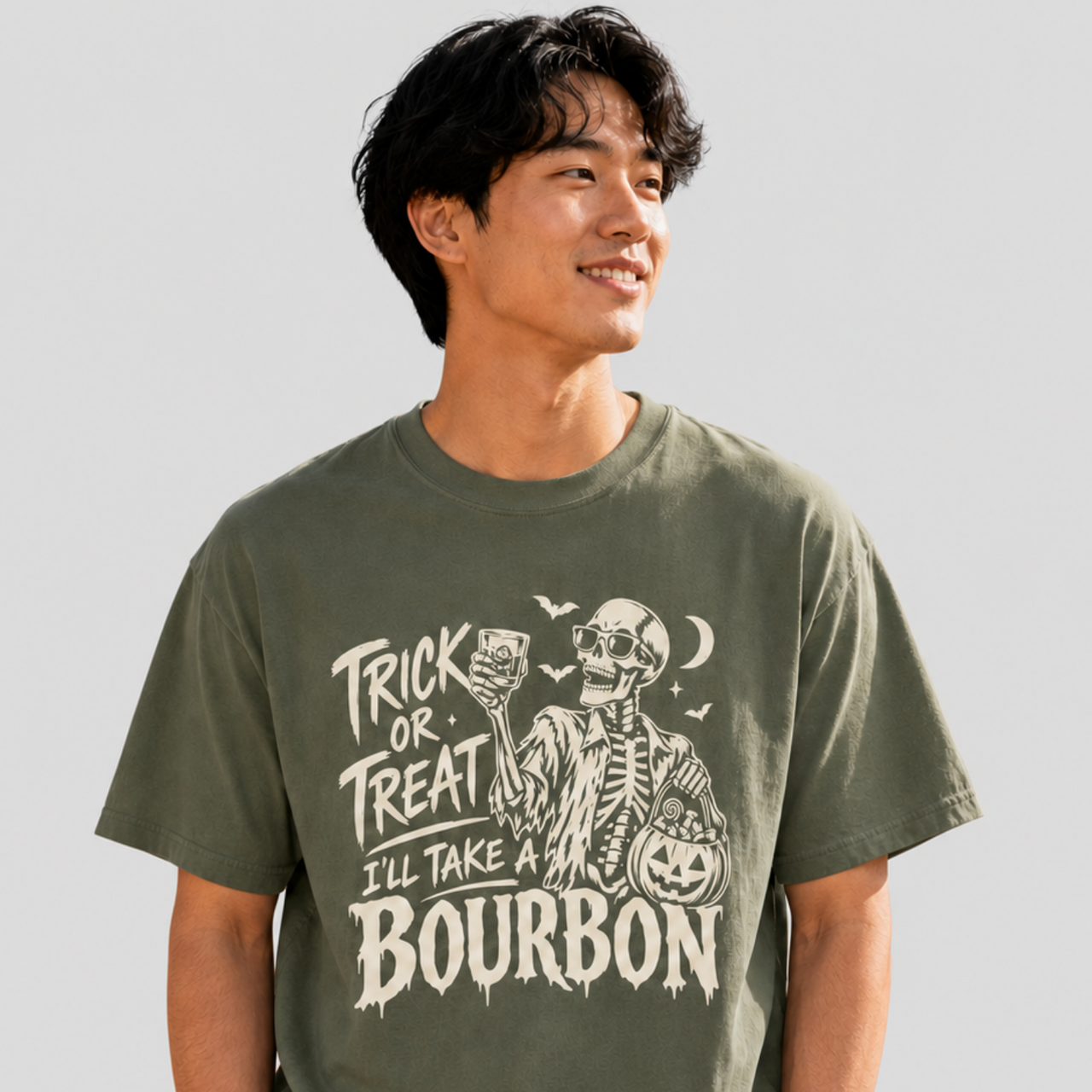 A man wearing an olive green graphic t-shirt with a white skeleton and bourbon-themed Halloween print, standing against a solid light gray background.