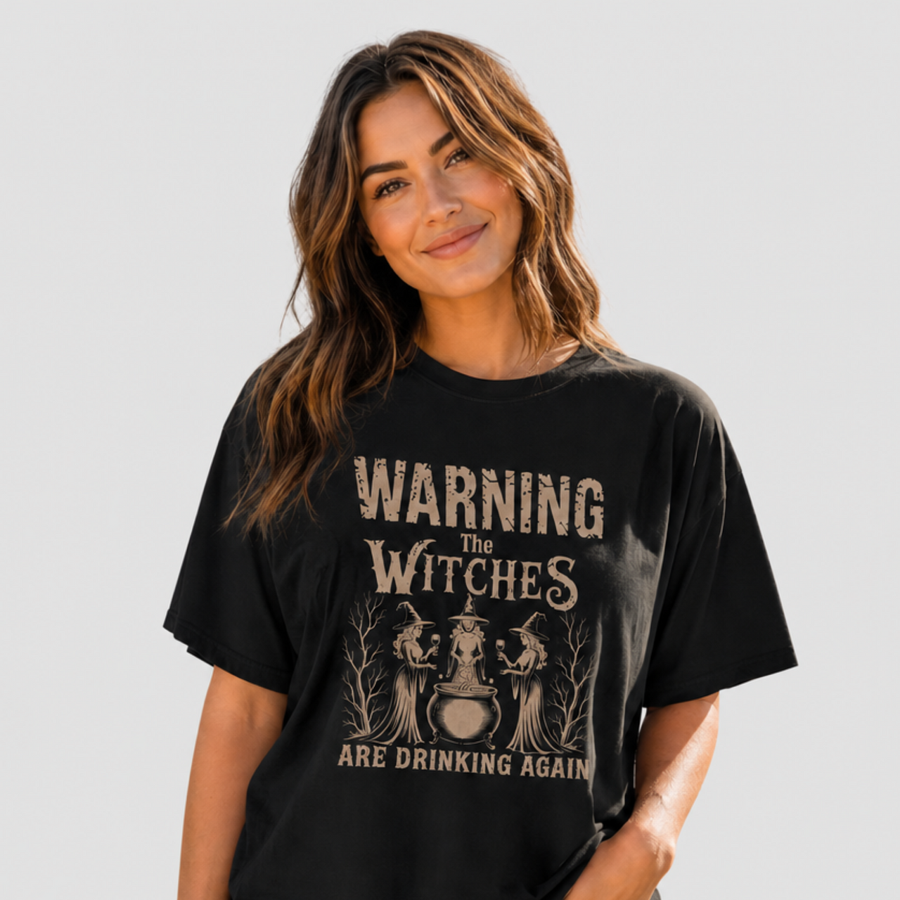 A woman wearing a black graphic t-shirt with a vintage-style beige print that reads 'WARNING THE WITCHES ARE DRINKING AGAIN' with illustrations of three witches and a cauldron. She is paired with light-wash blue jeans against a solid light gray background.
