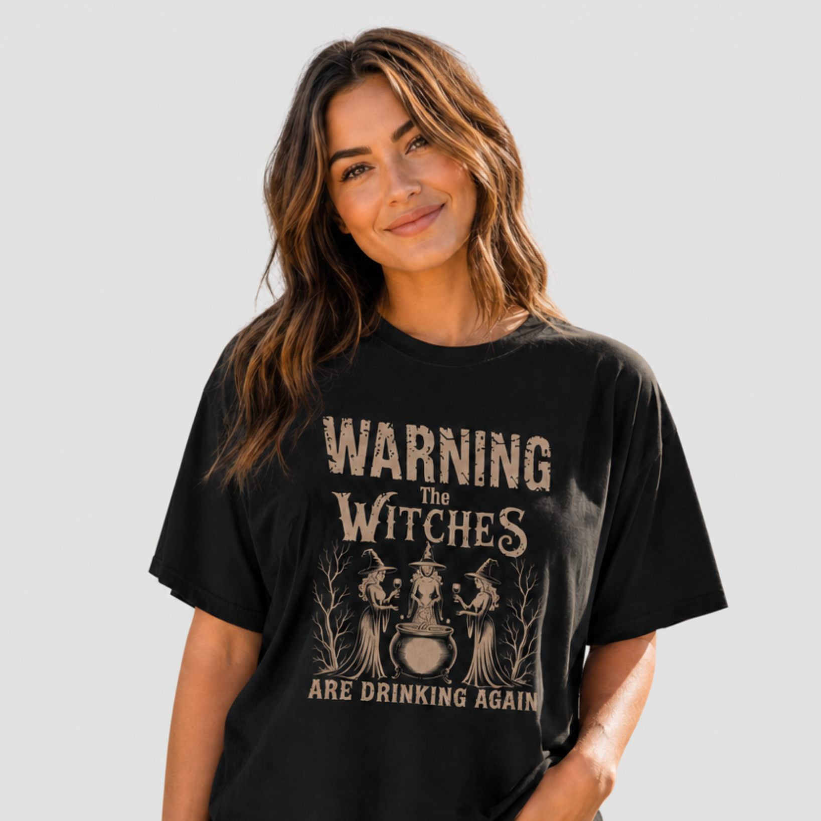A woman wearing a black graphic t-shirt with a vintage-style beige print that reads 'WARNING THE WITCHES ARE DRINKING AGAIN' with illustrations of three witches and a cauldron. She is paired with light-wash blue jeans against a solid light gray background.