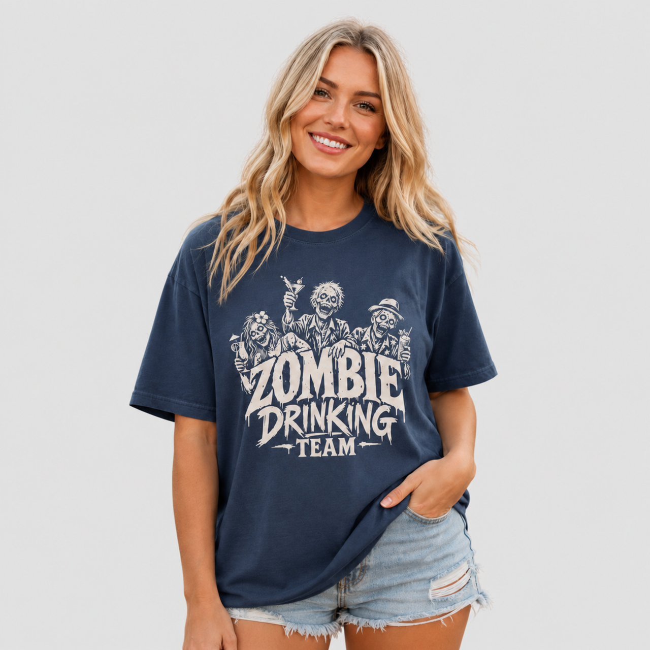 A woman modeling a navy blue graphic t-shirt with a distressed white print that reads 'ZOMBIE DRINKING TEAM' with cartoon zombies, paired with denim shorts, against a solid light gray background.