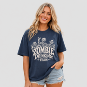 A woman modeling a navy blue graphic t-shirt with a distressed white print that reads 'ZOMBIE DRINKING TEAM' with cartoon zombies, paired with denim shorts, against a solid light gray background.