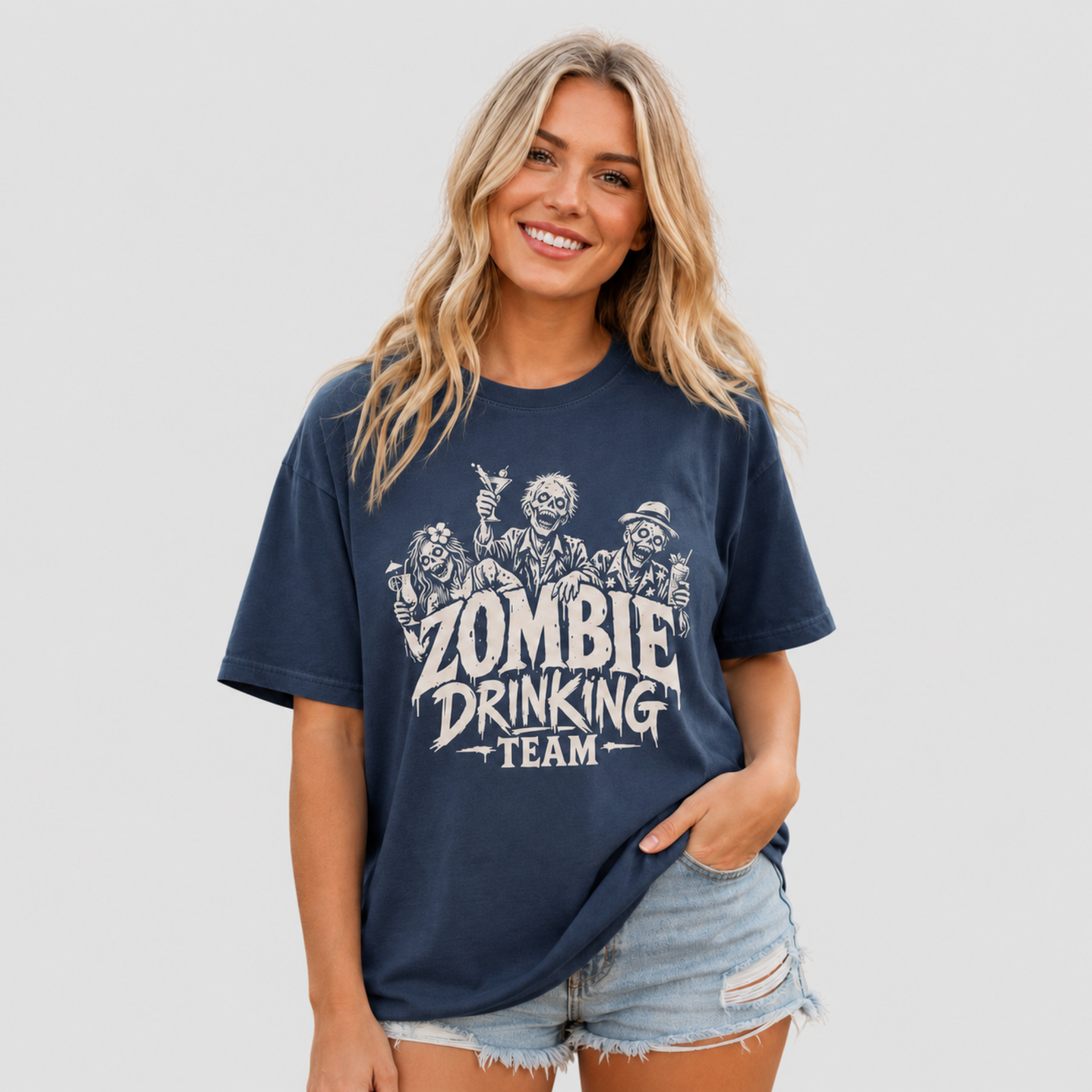 A woman modeling a navy blue graphic t-shirt with a distressed white print that reads 'ZOMBIE DRINKING TEAM' with cartoon zombies, paired with denim shorts, against a solid light gray background.