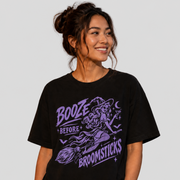 A woman wearing a black graphic t-shirt with a purple witch illustration and the text 'BOOZE BEFORE BROOMSTICKS', paired with light-wash jeans, set against a solid light gray background.