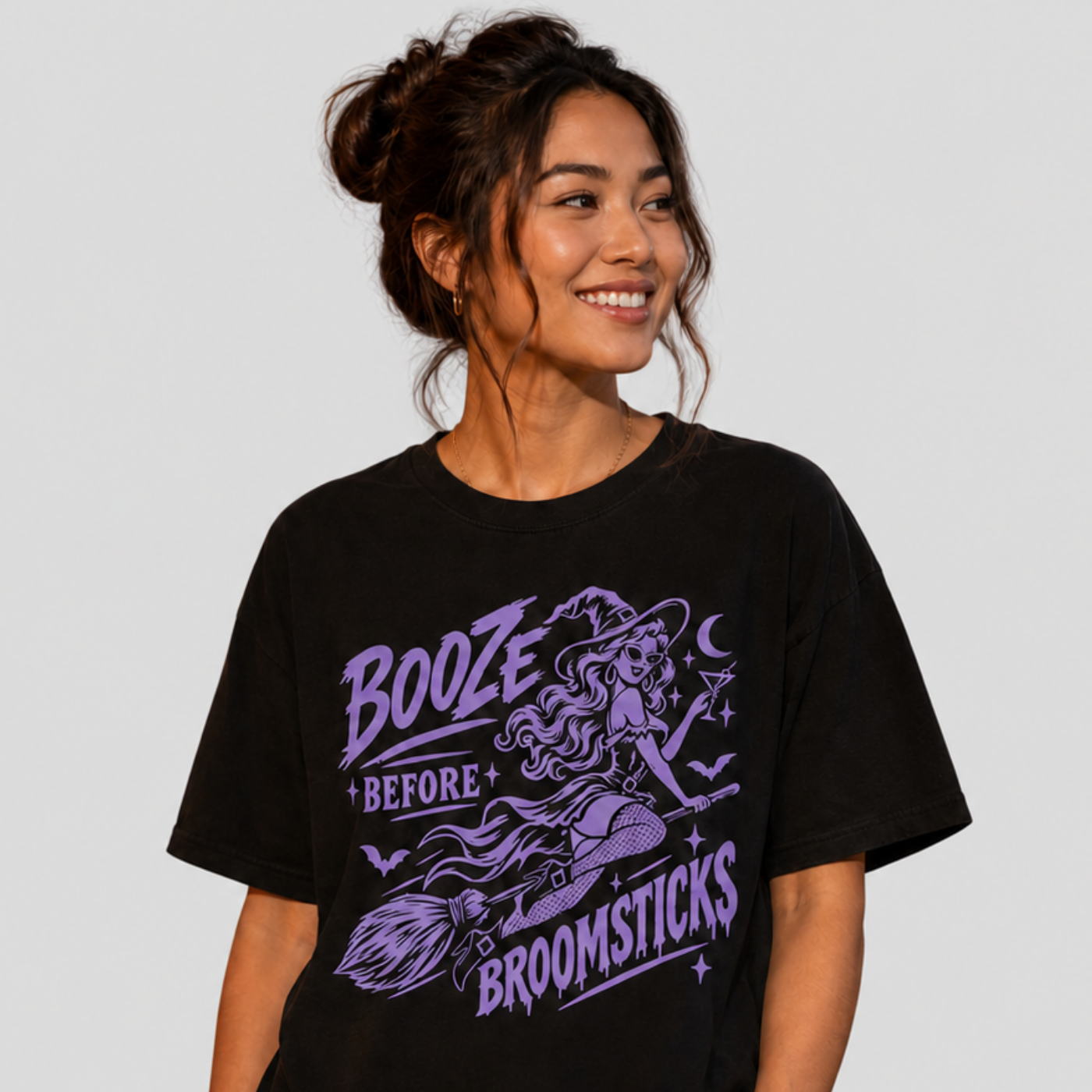 A woman wearing a black graphic t-shirt with a purple witch illustration and the text 'BOOZE BEFORE BROOMSTICKS', paired with light-wash jeans, set against a solid light gray background.