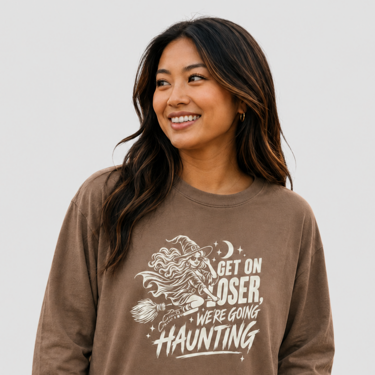 A woman wearing a brown long-sleeve graphic t-shirt with a white print that reads 'GET ON LOSER, WE'RE GOING HAUNTING' alongside a witch riding a broomstick, set against a plain light gray background.