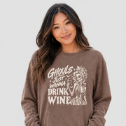 A woman wearing a brown long-sleeve graphic sweatshirt with a white skeleton and text that reads 'Ghouls Just Wanna Drink Wine,' paired with blue jeans, against a light gray studio background.
