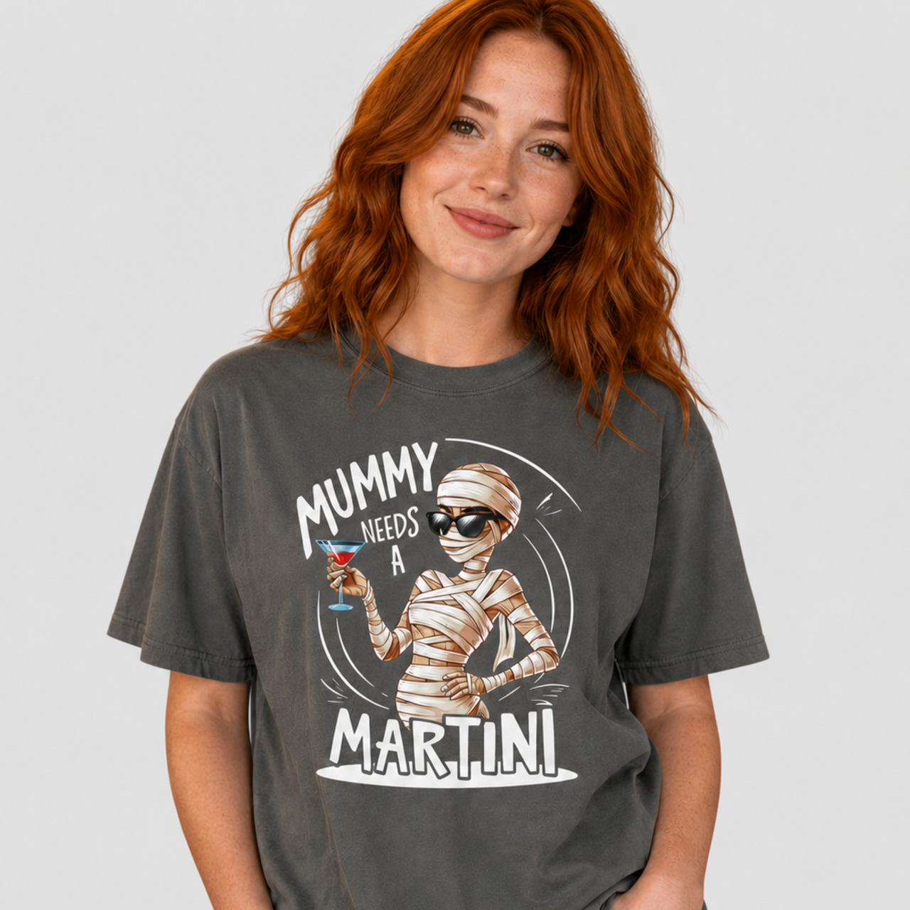 A woman with red hair wearing a dark gray graphic t-shirt that reads 'MUMMY NEEDS A MARTINI' with an illustration of a mummy holding a martini glass. She is also wearing light blue jeans, set against a solid light gray background.