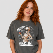 A woman with red hair wearing a dark gray graphic t-shirt that reads 'MUMMY NEEDS A MARTINI' with an illustration of a mummy holding a martini glass. She is also wearing light blue jeans, set against a solid light gray background.