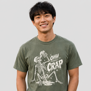 A smiling man wearing an olive green crewneck t-shirt with a white graphic of a skeleton drinking beer and the text 'OHH CRAP'.