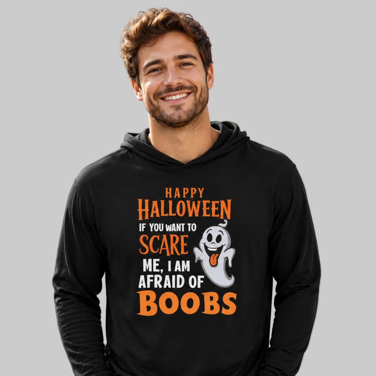A smiling man wearing a black long-sleeve hoodie with a Halloween graphic that reads 'Happy Halloween if you want to SCARE me, I am AFRAID of BOOBS' in orange and white, with a small ghost illustration, against a plain light gray background.