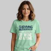 Woman wearing a light green t-shirt with the phrase 'Surviving Motherhood One Glass of Wine at a Time' funny Mother's day, beach vacation, spring break design