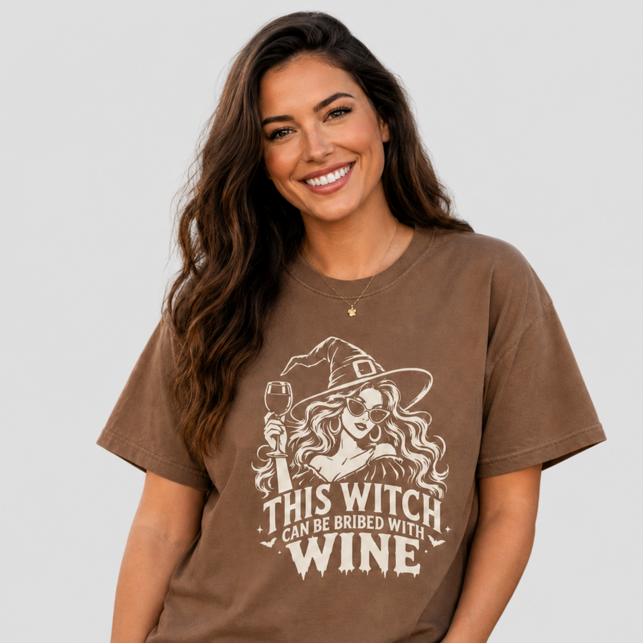 A woman wearing a brown graphic t-shirt with a white illustration of a witch holding a wine glass and the text 'THIS WITCH CAN BE BRIBED WITH WINE'.