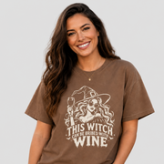 A woman wearing a brown graphic t-shirt with a white illustration of a witch holding a wine glass and the text 'THIS WITCH CAN BE BRIBED WITH WINE'.