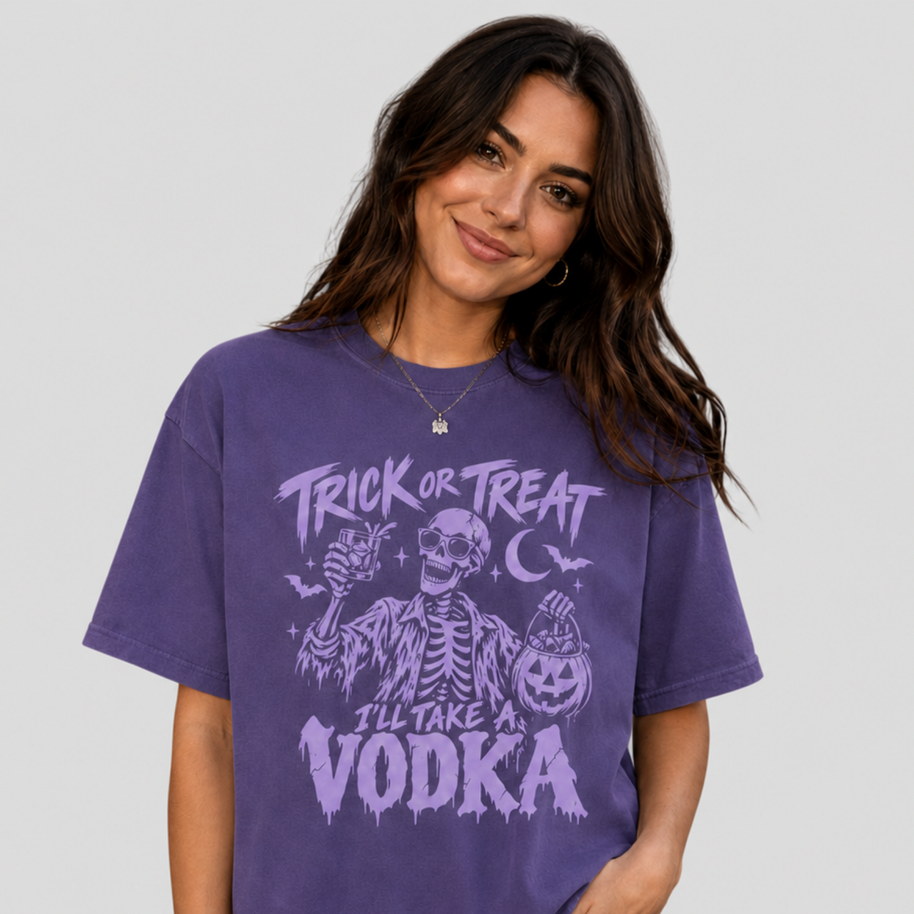 A woman wearing a purple short-sleeve graphic t-shirt with a 'Trick or Treat I'll Take a Vodka' skeleton and pumpkin design, paired with black pants, against a solid light gray background.