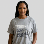 Woman wearing a gray t-shirt with the text 'Warning:the moms are drinking again' funny mother's day, beach trip, girls trip design
