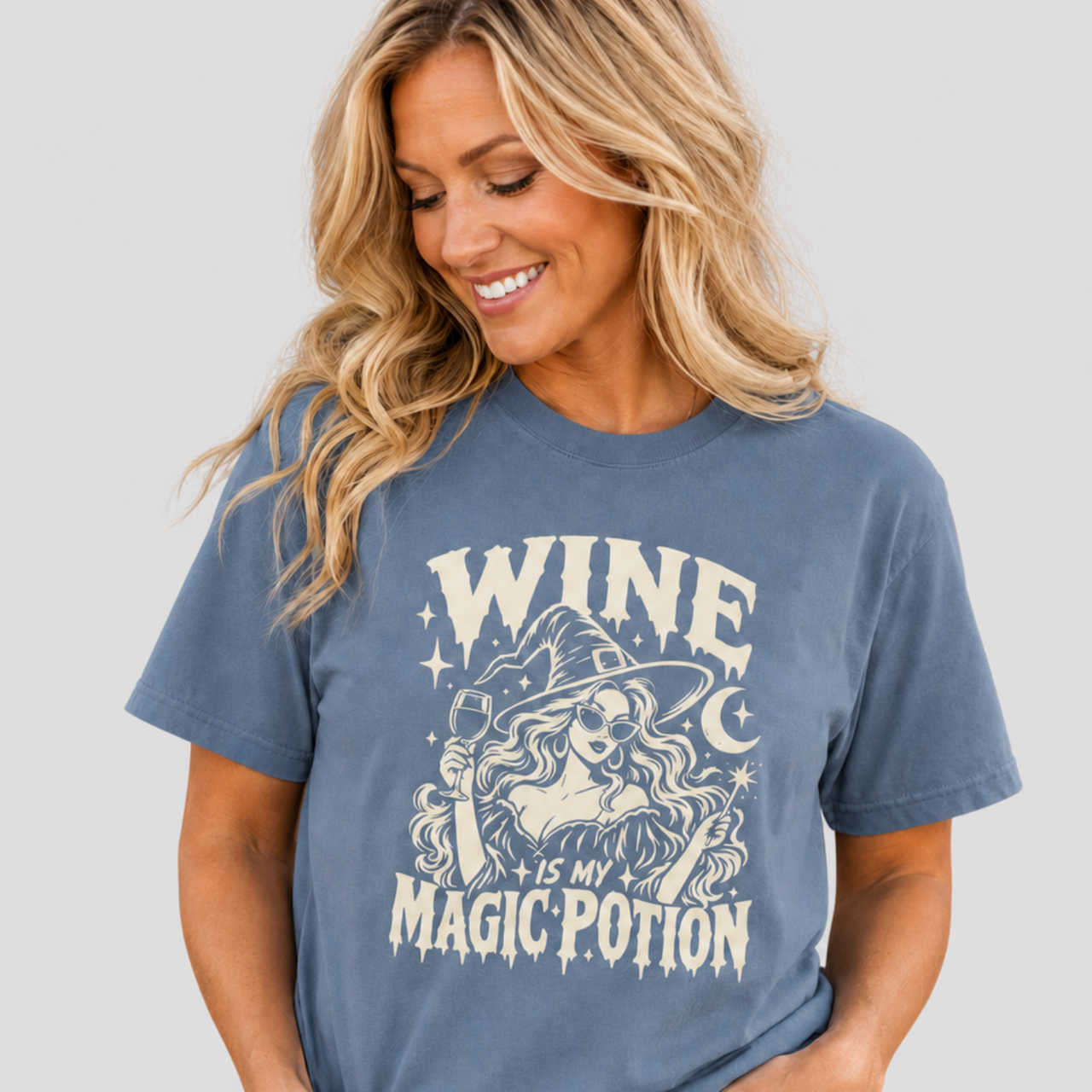 A woman wearing a slate blue graphic t-shirt with a cream-colored witch design and the text 'WINE IS MY MAGIC POTION', paired with light-wash blue jeans, against a solid light gray background.