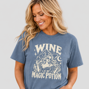 A woman wearing a slate blue graphic t-shirt with a cream-colored witch design and the text 'WINE IS MY MAGIC POTION', paired with light-wash blue jeans, against a solid light gray background.