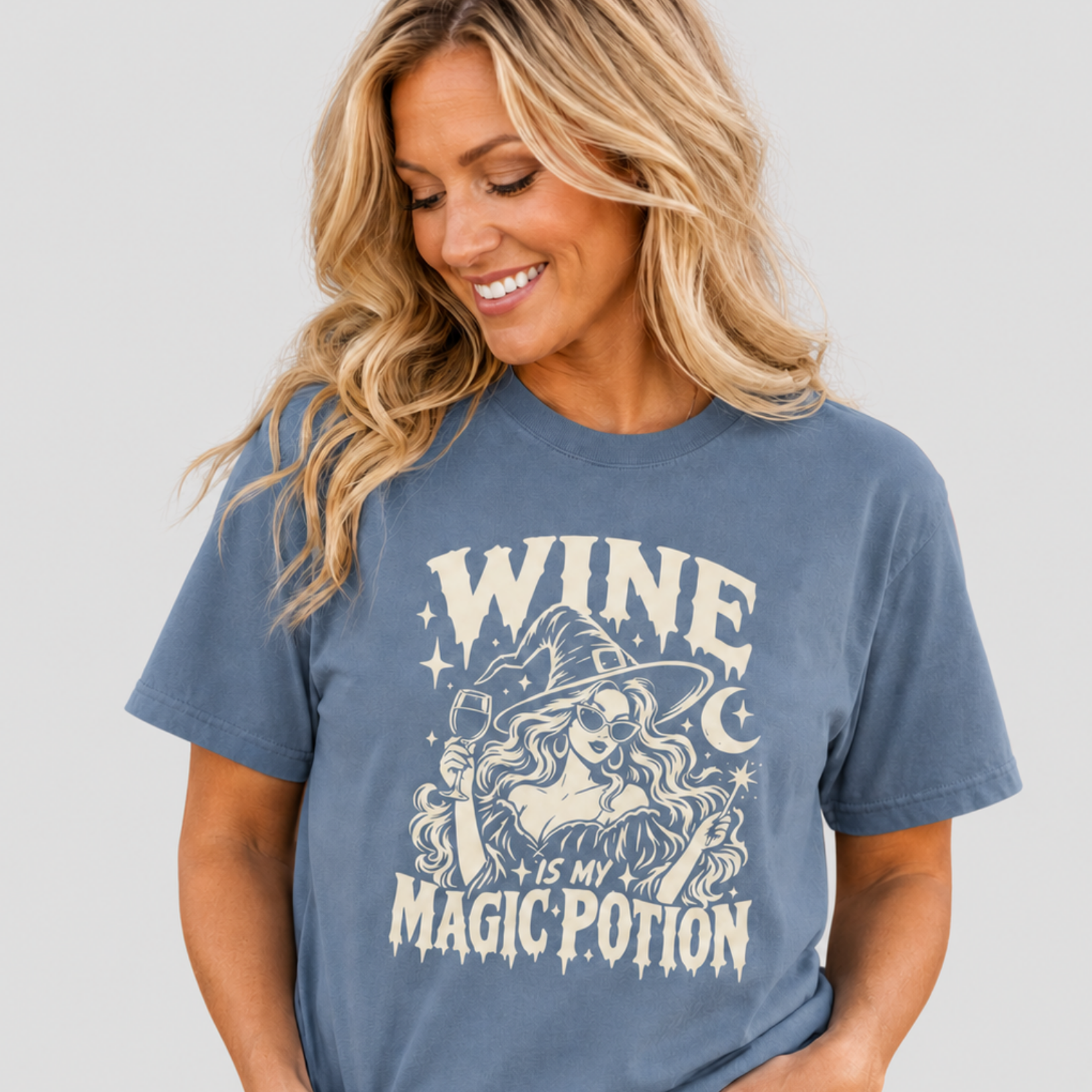 A woman wearing a slate blue graphic t-shirt with a cream-colored witch design and the text 'WINE IS MY MAGIC POTION', paired with light-wash blue jeans, against a solid light gray background.